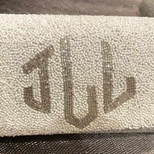 May include: A white beaded clutch bag with a silver monogram of the letters "JUL".