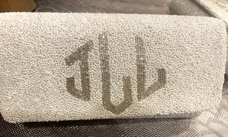 May include: A white beaded clutch bag with a silver monogram of the letters "JUL".