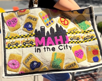 May include: A large off-white tote bag featuring intricate beadwork with a New York City theme. The design includes a black skyline, yellow taxi cabs, the text "MAHJ in the City," and various playing card motifs with "JOKER" and "NYC" labels.