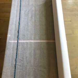 May include: A large roll of white fabric next to a flat, unfolded piece of the same material. A blue measuring tape is laid out along the length of the fabric, showing measurements in inches. The fabric appears to be a light, translucent material.