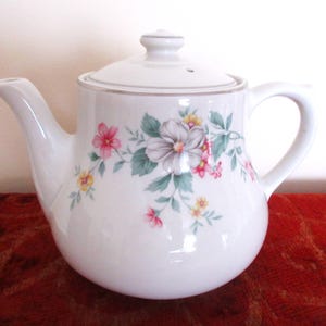May include: White ceramic teapot with a floral design featuring pink, white, and yellow flowers. The teapot has a lid and a handle.