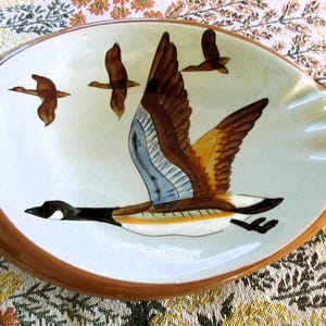 May include: Oval ceramic ashtray with a brown rim and a white interior. The interior features a hand-painted design of Canada geese in flight, with brown, blue, and yellow details. The ashtray has a classic design.