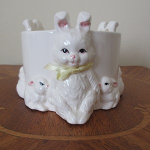 May include: White ceramic planter shaped like a bunny rabbit with two smaller bunnies at its feet. The bunny has a yellow ribbon around its neck.