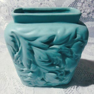 May include: A turquoise ceramic vase with a rectangular opening and a rounded body. The vase features a raised leaf design that covers the entire surface. The vase is approximately 7 inches tall and 6 inches wide.