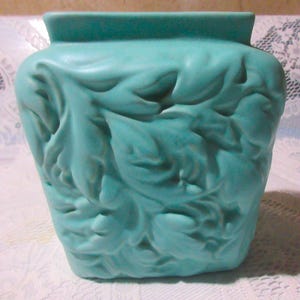 May include: A turquoise ceramic vase with a square base and a rectangular top. The vase features a raised floral design covering its surface. The color is a muted, seafoam green.