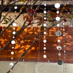 May include: Outdoor scene featuring a tree with colorful autumn leaves and decorative hanging ornaments. The ornaments are round, reflective, and strung together, creating a shimmering effect. A wooden wall and brick structure form the backdrop.