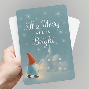 May include: A Christmas card held by a hand, featuring a whimsical illustration. The card has a blue background with the text "All is Merry, All is Bright". A gnome with a red hat and blue coat stands in front of snow-covered houses.