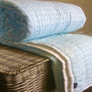 May include: A light blue and white patterned quilt with a white border. The quilt is folded and placed on a wicker basket.
