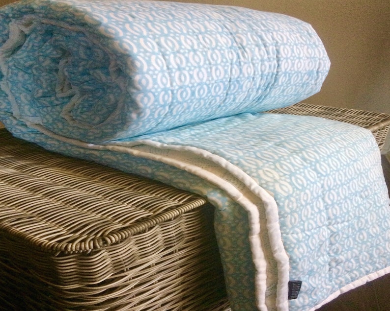 May include: A light blue and white patterned quilt with a white border. The quilt is folded and placed on a wicker basket.