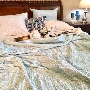 May include: A white teapot and a white candle sit on a wooden tray on a bed with a light blue patterned quilt. The bed has white pillows and a light blue patterned pillow.