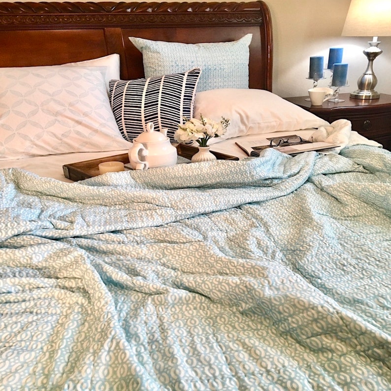 May include: A white teapot and a white candle sit on a wooden tray on a bed with a light blue patterned quilt. The bed has white pillows and a light blue patterned pillow.