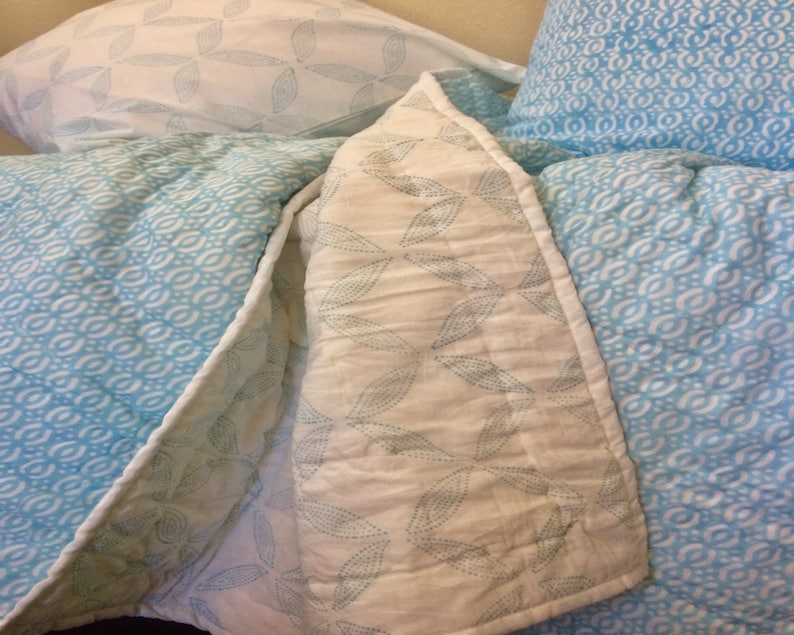 May include: A white and blue patterned quilt with a white and blue patterned pillow on top. The quilt has a leaf design and the pillow has a wave design.
