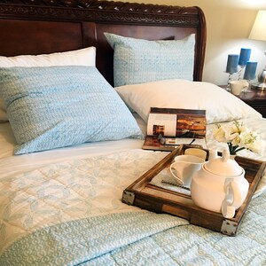 May include: A white teapot and a white cup on a wooden tray with a magazine and white flowers on a bed with blue and white bedding.