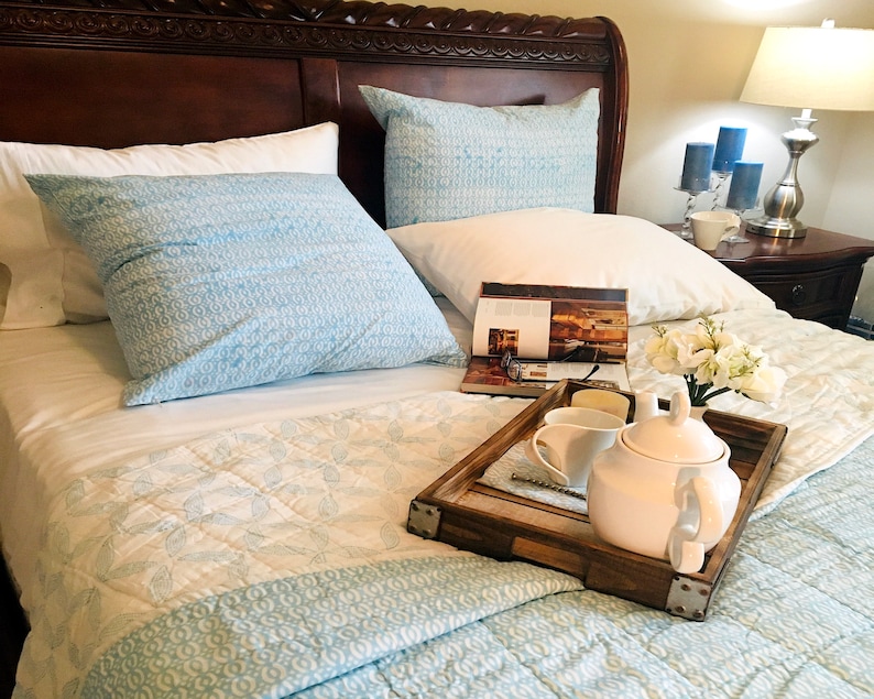 May include: A white teapot and a white cup on a wooden tray with a magazine and white flowers on a bed with blue and white bedding.