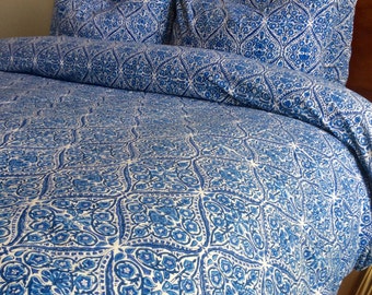 May include: A bed with a blue and white patterned comforter and matching pillow shams. The bedding features a repeating floral and geometric design. The headboard is made of dark wood.