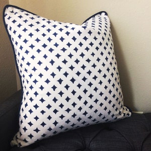 May include: A white pillow with a navy blue diamond pattern. The pillow has a navy blue border and is sitting on a dark gray couch.
