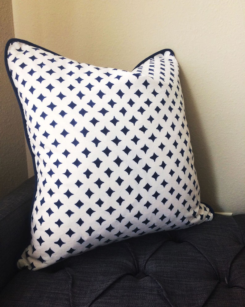 May include: A white pillow with a navy blue diamond pattern. The pillow has a navy blue border and is sitting on a dark gray couch.