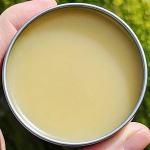 Puede incluir: Una lata redonda de metal llena de un bálsamo amarillo pálido.