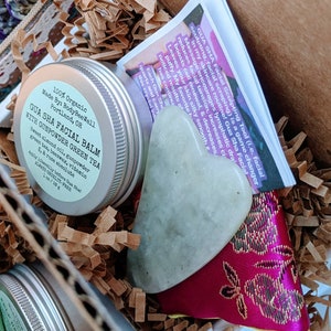 Puede incluir: Un set de regalo de belleza que contiene un bálsamo facial Gua Sha, una herramienta Gua Sha de jade y un folleto. El bálsamo está en una lata plateada con una etiqueta verde. La herramienta Gua Sha es verde claro y en forma de corazón.