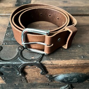 May include: A tan leather belt coiled into a circle, featuring a polished silver-tone metal buckle with a single prong and a matching leather keeper loop. The belt strap has a smooth texture and visible pre-punched holes for adjustment.