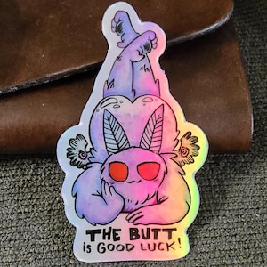May include: A colorful sticker featuring a cartoon moth-like creature with red eyes and a rainbow gradient. The text "THE BUTT is GOOD LUCK!" is printed below the creature.