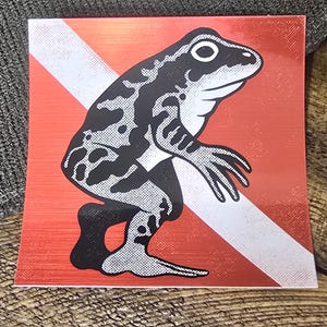 May include: A square sticker featuring a black and white illustration of a frog standing upright in a human-like pose. The frog is positioned over a red background with a diagonal white stripe, mimicking the design of a diver down flag.