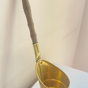 May include: A brass ladle with a wooden handle. The ladle has a rounded bowl and a long, slender handle. The handle is attached to the bowl with a metal hinge.