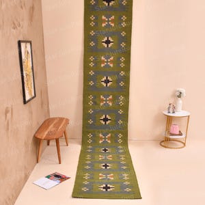 May include: A long, rectangular green rug with a geometric pattern of gray, black, and tan squares and crosses. The rug is made of a woven material and is displayed on a light-colored floor. The rug is approximately 8 feet long.