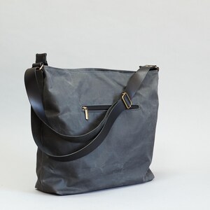 May include: A charcoal gray shoulder bag featuring a textured, matte finish and a black adjustable strap with brass-toned hardware. The exterior includes a horizontal zippered pocket with a metallic pull, providing a functional design for daily use.