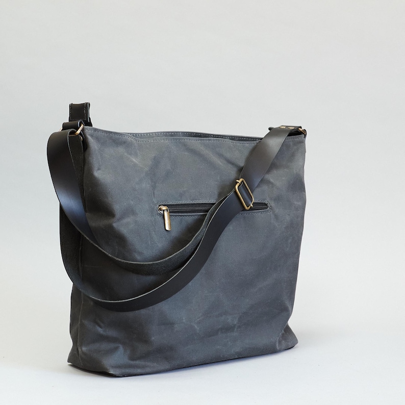 May include: A charcoal gray shoulder bag featuring a textured, matte finish and a black adjustable strap with brass-toned hardware. The exterior includes a horizontal zippered pocket with a metallic pull, providing a functional design for daily use.