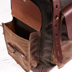 Puede incluir: Una mochila de cuero marr&oacute;n y lona con m&uacute;ltiples bolsillos, incluyendo un bolsillo grande con solapa y cierre de cuero y un bolsillo m&aacute;s peque&ntilde;o con un bot&oacute;n de metal.