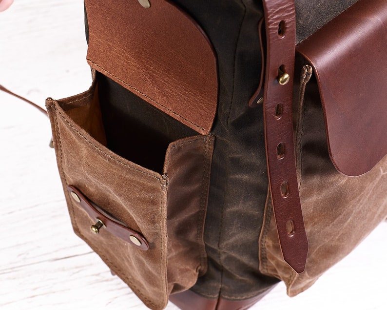 Puede incluir: Una mochila de cuero marr&oacute;n y lona con m&uacute;ltiples bolsillos, incluyendo un bolsillo grande con solapa y cierre de cuero y un bolsillo m&aacute;s peque&ntilde;o con un bot&oacute;n de metal.