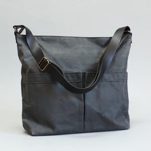 May include: A charcoal gray crossbody bag made of a durable, matte fabric. The bag features a long, adjustable black strap with a brass-toned metal buckle and two large, vertical-stitched exterior pockets on the front for storage and organization.
