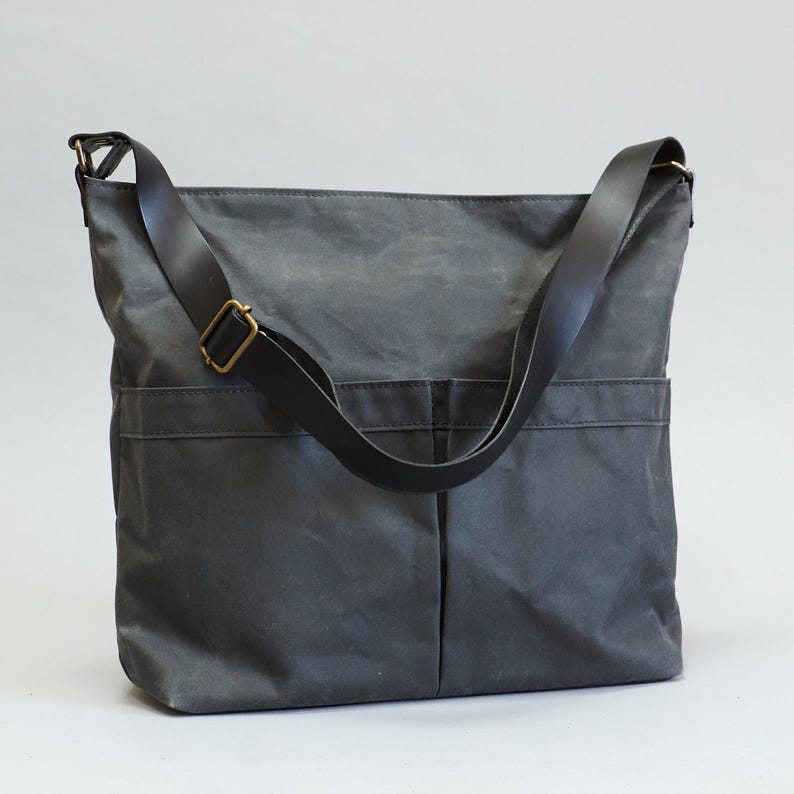 May include: A charcoal gray crossbody bag made of a durable, matte fabric. The bag features a long, adjustable black strap with a brass-toned metal buckle and two large, vertical-stitched exterior pockets on the front for storage and organization.