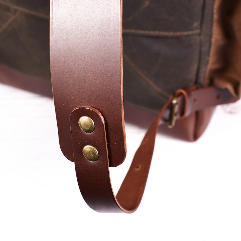 Puede incluir: Primer plano de una correa de mochila de cuero marr&oacute;n con dos remaches de lat&oacute;n. La correa es ajustable y tiene una hebilla.