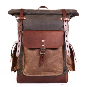 Puede incluir: Una mochila de lona marr&oacute;n y verde con correas y hebillas de cuero. La mochila tiene un compartimento principal grande y dos bolsillos m&aacute;s peque&ntilde;os en la parte delantera.