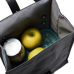 Puede incluir: Una bolsa de almuerzo negra con interior gris, que contiene dos manzanas verdes, un recipiente peque&ntilde;o y un tarro con tapa estampada en rojo y blanco. La bolsa tiene un asa negra y est&aacute; abierta, mostrando el contenido.