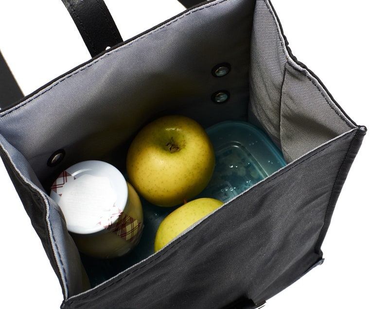 Puede incluir: Una bolsa de almuerzo negra con interior gris, que contiene dos manzanas verdes, un recipiente peque&ntilde;o y un tarro con tapa estampada en rojo y blanco. La bolsa tiene un asa negra y est&aacute; abierta, mostrando el contenido.