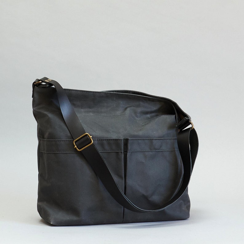 May include: A dark charcoal gray crossbody bag featuring a rectangular silhouette, two large front exterior pockets, and a wide adjustable shoulder strap. The strap is secured with a metallic brass-toned rectangular buckle and matching hardware.