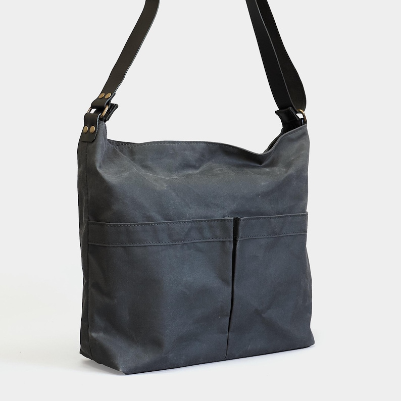 May include: A charcoal gray waxed canvas shoulder bag featuring a spacious main compartment, two front exterior pockets, and a wide adjustable shoulder strap attached with brass-toned metal rivets and hardware for a secure and functional design.
