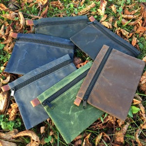 May include: Five small zippered pouches made of waxed canvas in different colors: dark blue, black, brown, green, and light blue. The pouches are lying on a bed of fallen leaves and grass.