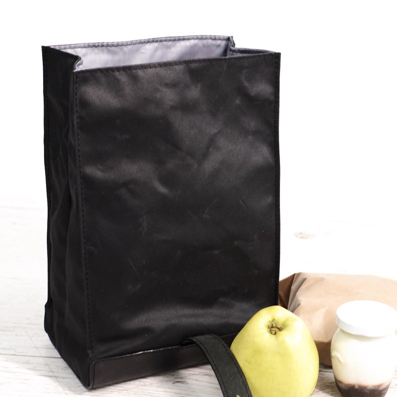 Puede incluir: Una bolsa de almuerzo negra con asa, hecha de un material brillante. La bolsa est&aacute; junto a una manzana verde, un s&aacute;ndwich envuelto y un peque&ntilde;o recipiente de yogur.