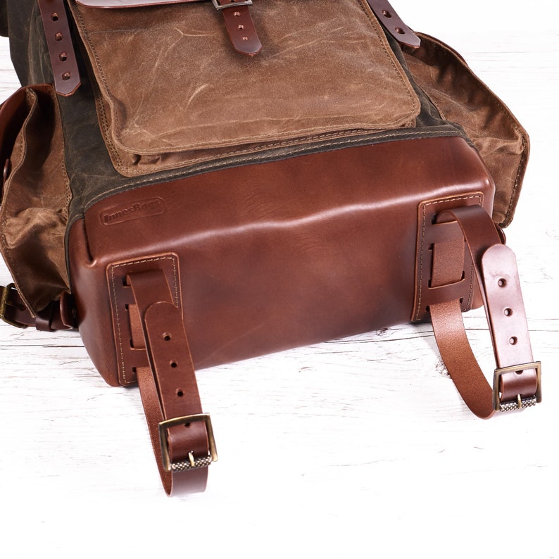 Puede incluir: Una mochila de cuero marr&oacute;n con una solapa de lona y una base de lona verde. La mochila tiene dos correas ajustables y un asa de cuero.