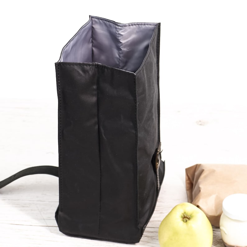 Puede incluir: Bolsa de almuerzo negra aislada con interior gris. La bolsa tiene una correa para el hombro y un cierre de hebilla. Una manzana, un tarro con tapa blanca y una bolsa de papel marr&oacute;n est&aacute;n en el fondo.