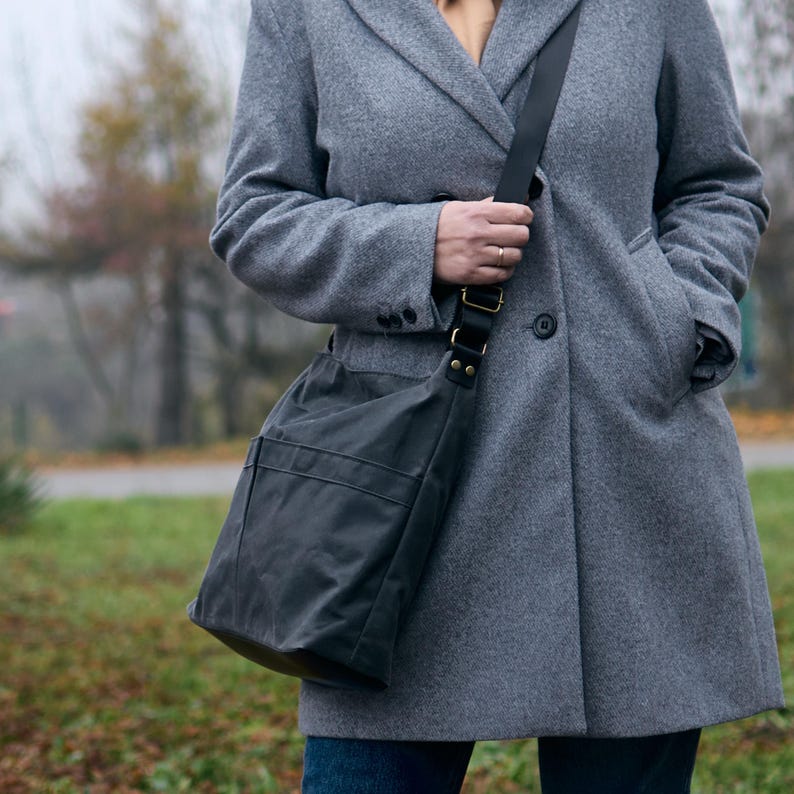 May include: A charcoal gray crossbody bag with a smooth, matte finish and a wide black adjustable strap. The bag features a vertical seam detail on the front panel and is secured with black metal hardware, including rivets and a rectangular buckle.