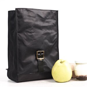 Puede incluir: Mochila negra de forma rectangular, con cierre de hebilla y base de cuero. La bolsa est&aacute; hecha de un material brillante y arrugado. Una pera amarilla y un peque&ntilde;o tarro de yogur est&aacute;n al lado de la bolsa.