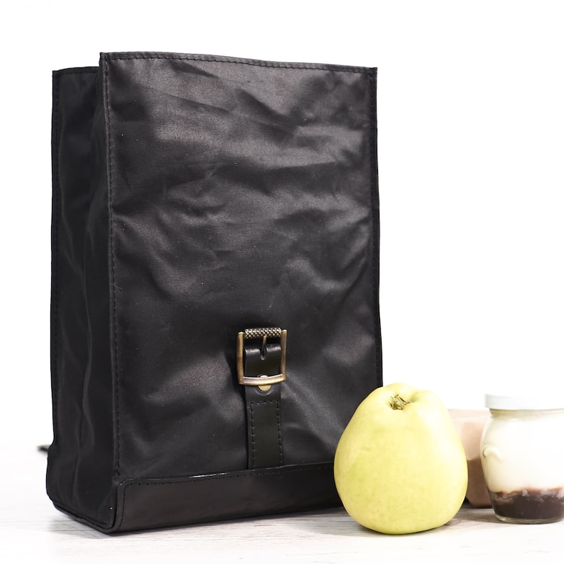 Puede incluir: Mochila negra de forma rectangular, con cierre de hebilla y base de cuero. La bolsa est&aacute; hecha de un material brillante y arrugado. Una pera amarilla y un peque&ntilde;o tarro de yogur est&aacute;n al lado de la bolsa.