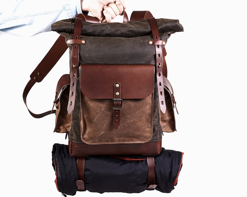 Puede incluir: Una mochila de lona marr&oacute;n y verde con detalles y correas de cuero. La mochila tiene un cierre de rollo y un bolsillo frontal grande. Est&aacute; siendo sostenida por una mano.