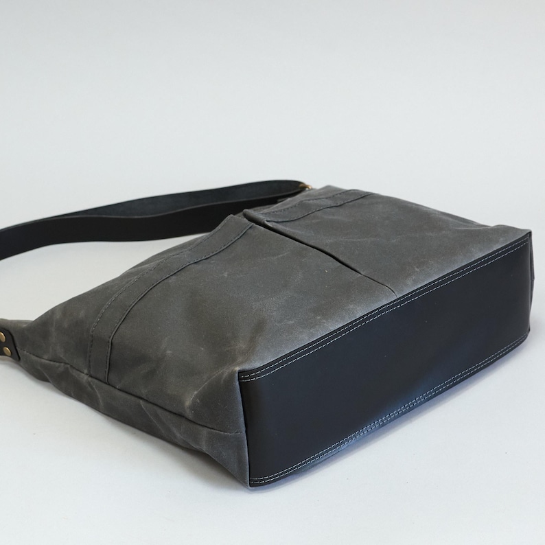 May include: A charcoal gray waxed canvas shoulder bag featuring a solid black leather base and a matching black leather shoulder strap. The bag has a structured design with visible stitching along the bottom panel and metal rivets at the strap attachment.
