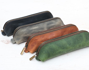 May include: Four leather pencil cases in different colors: black, gray, brown, and green. Each case has a zipper closure.